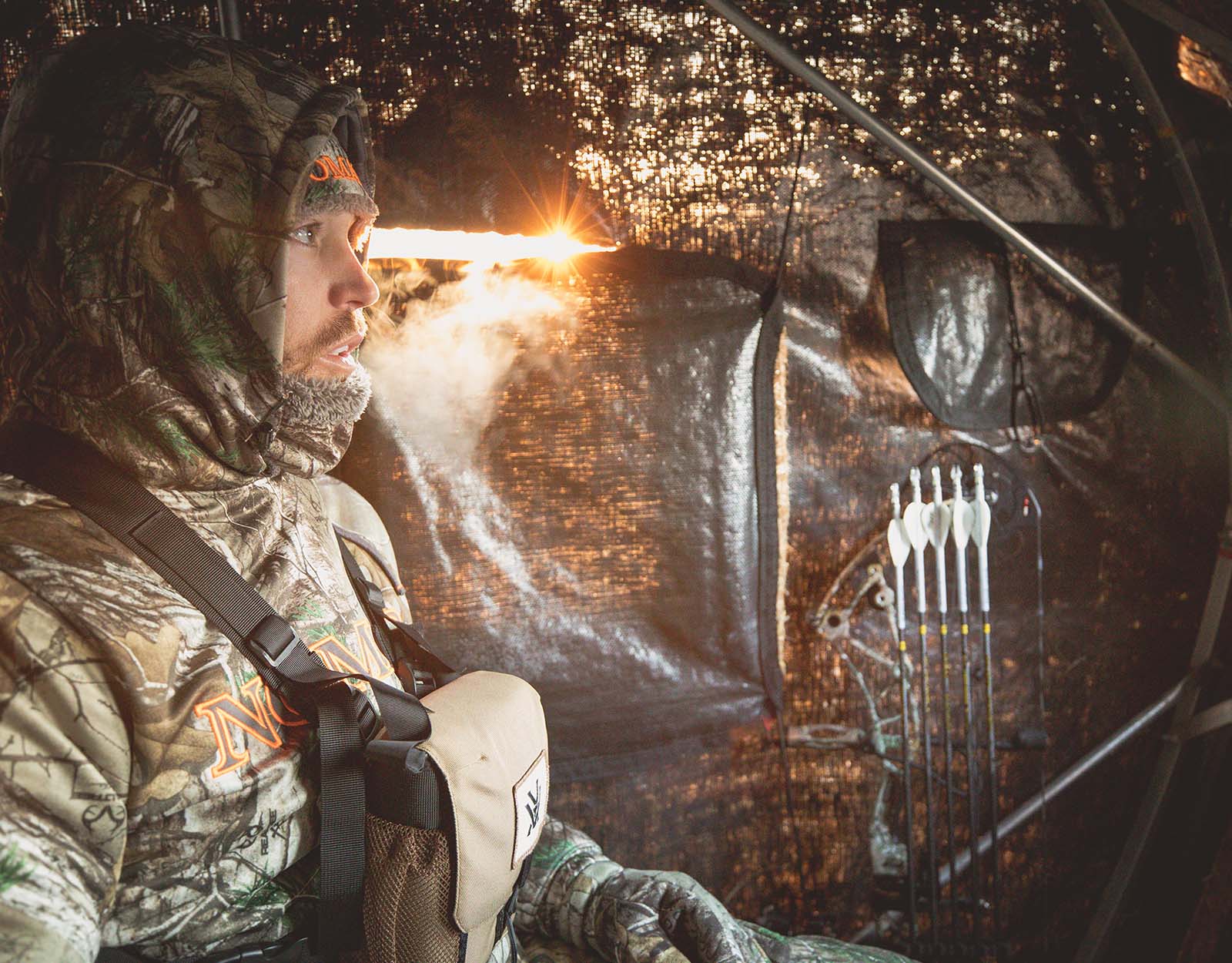 A bowhunter wearing cold weather gear sits in deer blind.